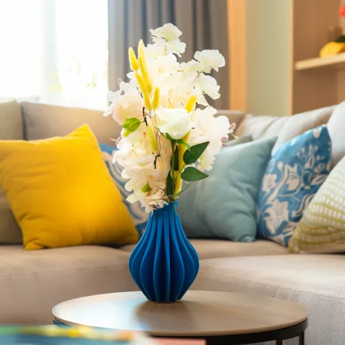 Blauwe vaas met bloemen op een ronde houten tafel in een woonkamer, omringd door kleurrijke kussens op een beige bank.