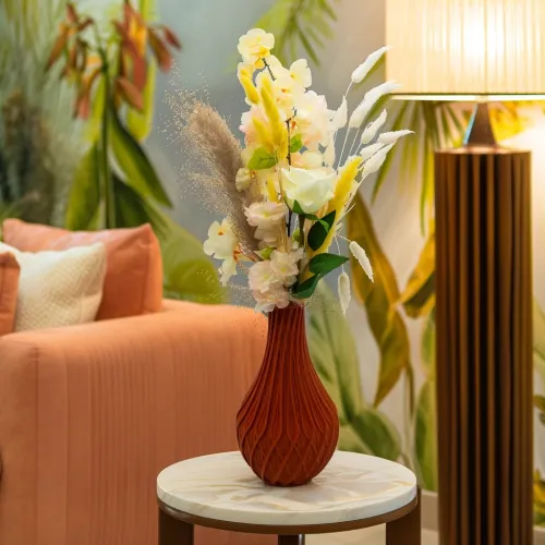 Een oranje vaas met kleurrijke bloemen staat op een ronde tafel. Op de achtergrond is een zachte, zalmroze fauteuil, een vloerlamp en een wand met bladeren zichtbaar.