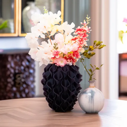 Een decoratieve zwarte vaas met gestructureerd patroon, gevuld met witte en roze bloemen, naast een kleine ronde vaas met groene takken, op een houten tafel.