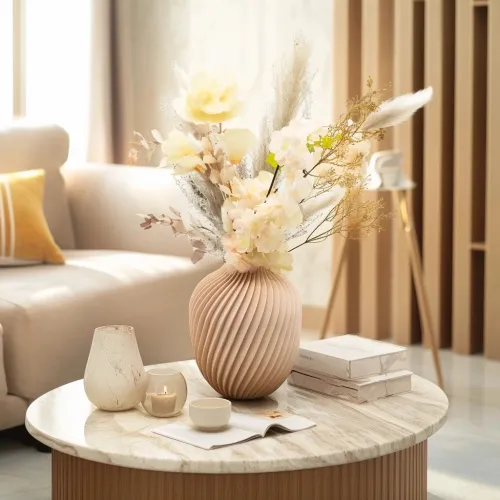 Een beige keramische vaas met bloemenarrangement op een ronde marmeren salontafel, omringd door een kaars, opengeslagen boek en koffiekopje, in een lichte woonkamer.
