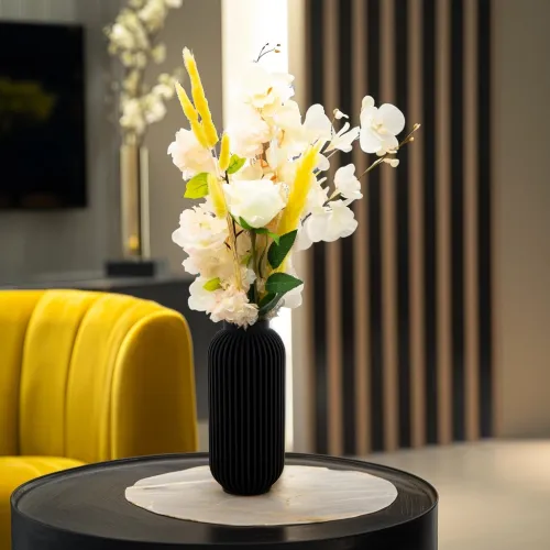 Een zwarte vaas met witte en gele bloemen op een ronde tafel, naast een gele stoel in een moderne ruimte.