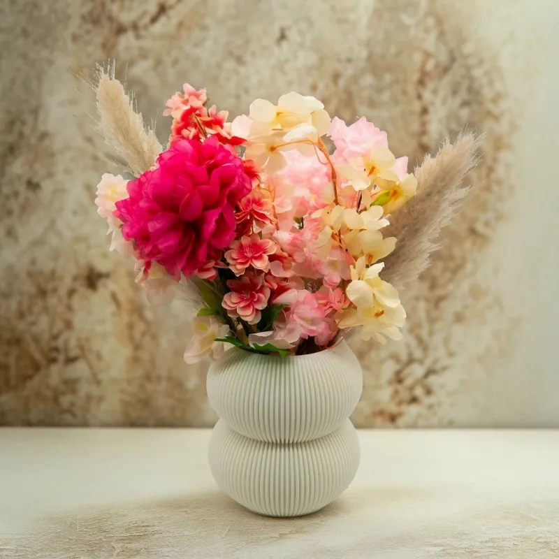 Een vaas met kleurrijke bloemenarrangement, waaronder roze en rode bloemen, tegen een neutrale achtergrond.