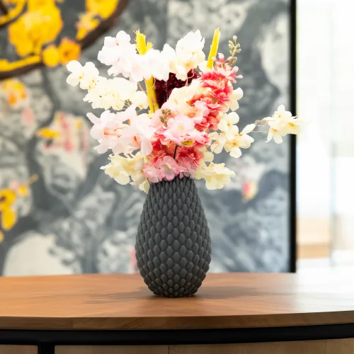 Grijze vaas met roze en witte bloemen op een houten tafel, tegen een grijze achtergrond met gele details.